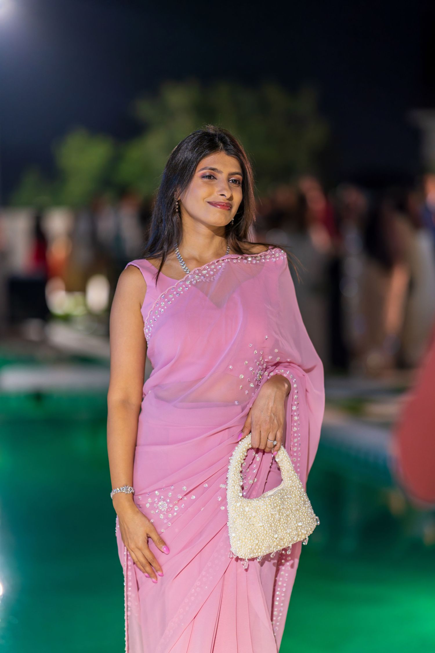 Woman in pink embellished sari poses elegantly by pool at night event with cream handbag.