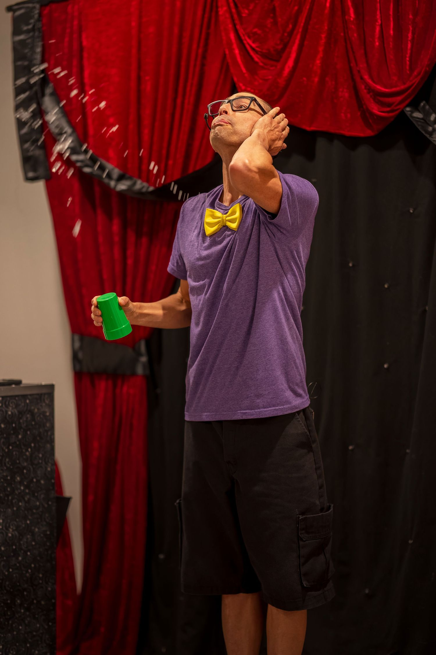 Magician in purple shirt performing liquid trick on stage with red curtain.