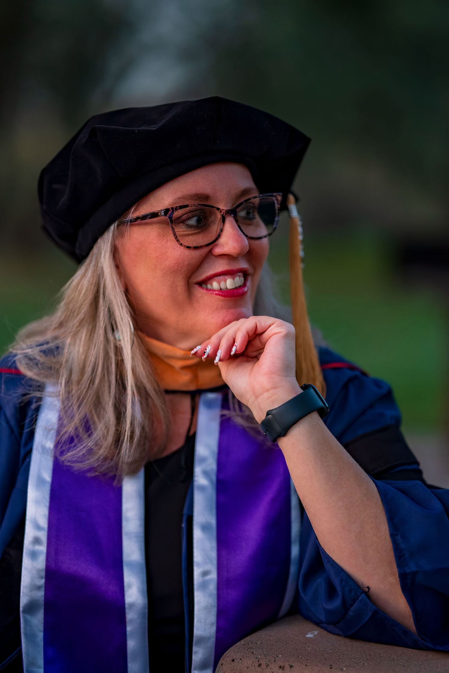 Graduate in academic regalia and cap smiles while posing against blurred natural background.