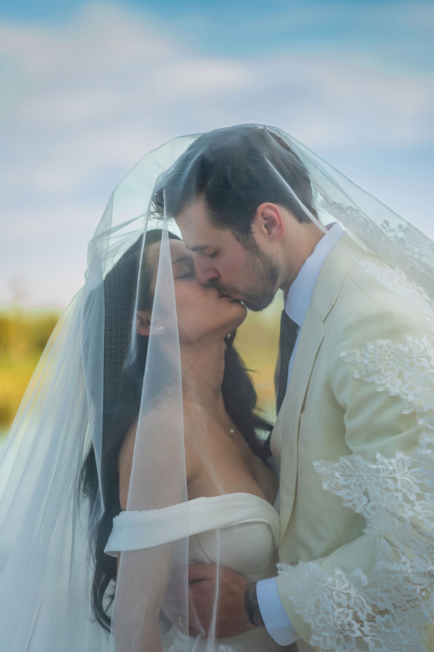 A romantic wedding kiss captured through a delicate lace veil against a soft sky background.