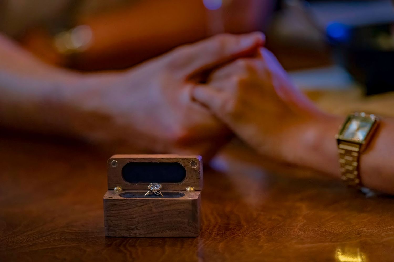 A wooden ring box sits on a table with hands and jewelry visible in soft lighting.