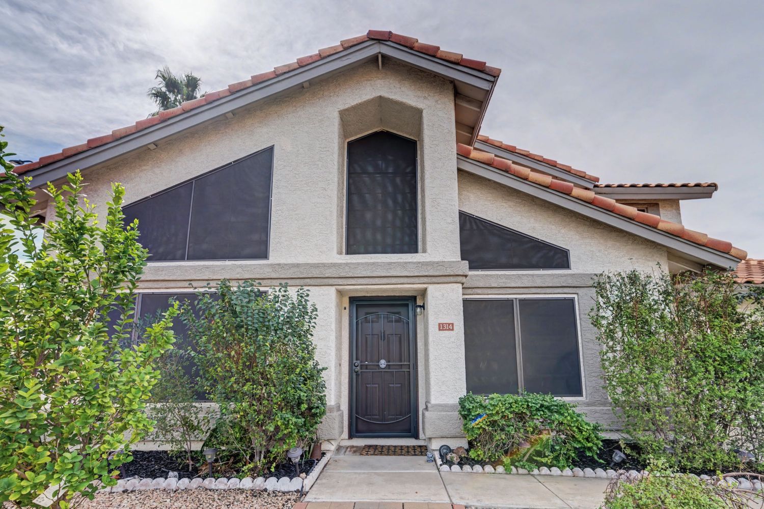 Two-story stucco home with red tile roof, dark screened windows, arched entryway, and lush landscaping in a sunny neighborhood.