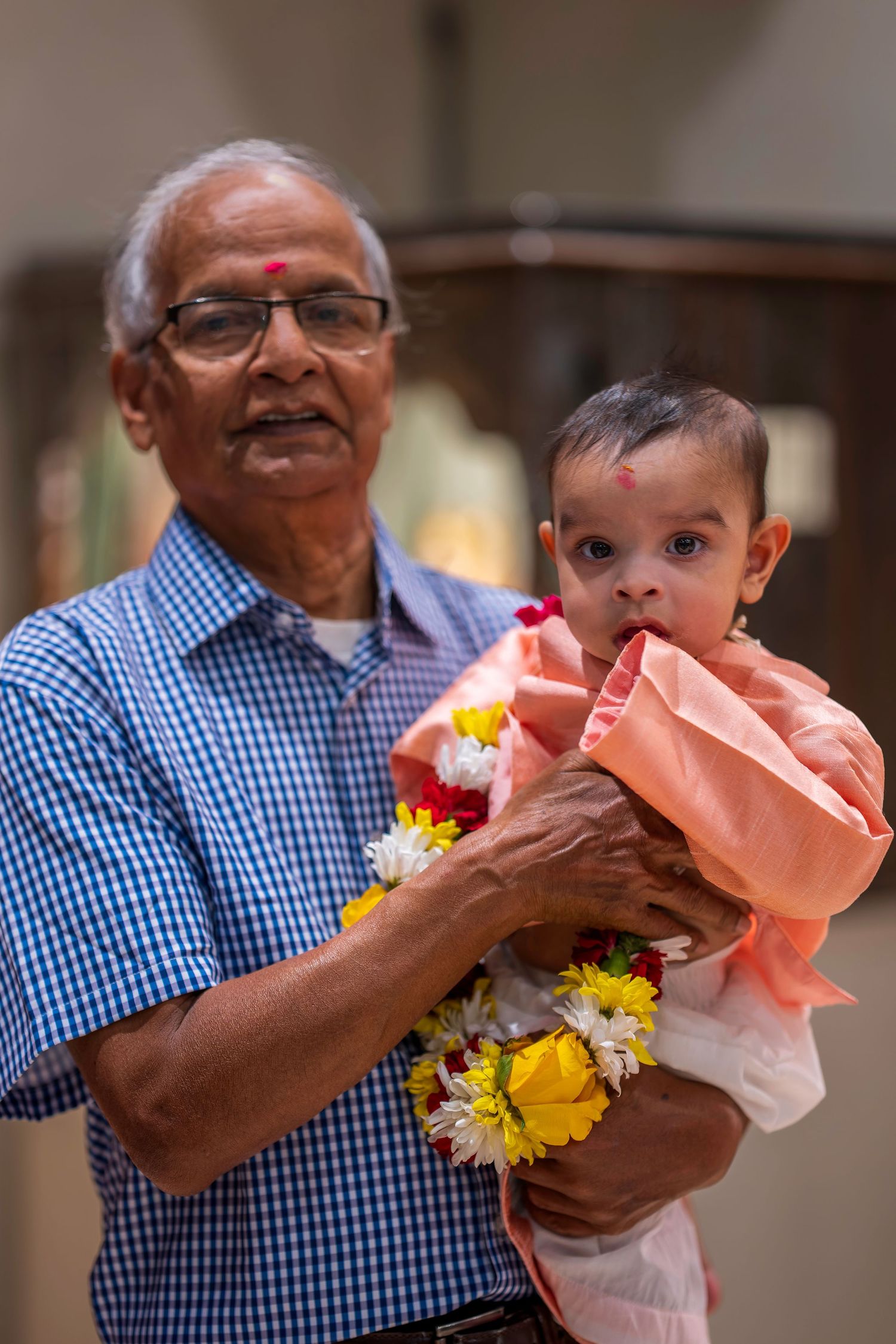 Elderly Indian man holding a baby adorned with flower garland and red tilak during a traditional ceremony.