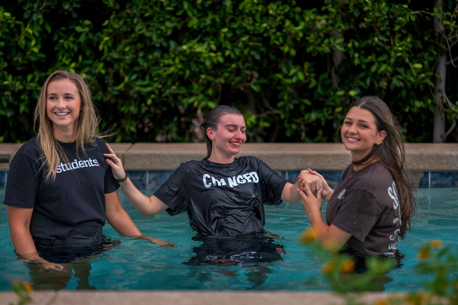 Three young women smiling in a pool during a baptism ceremony, wearing black shirts.