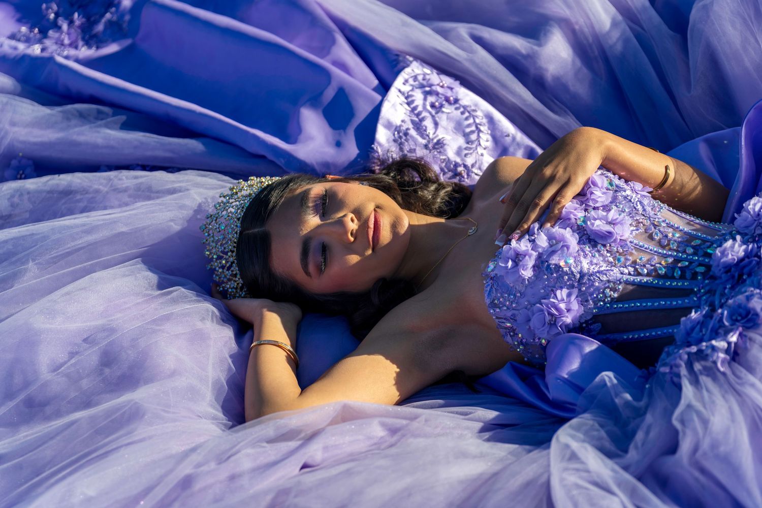 Young woman in lavender ball gown with floral details and tiara lying gracefully on purple tulle fabric.