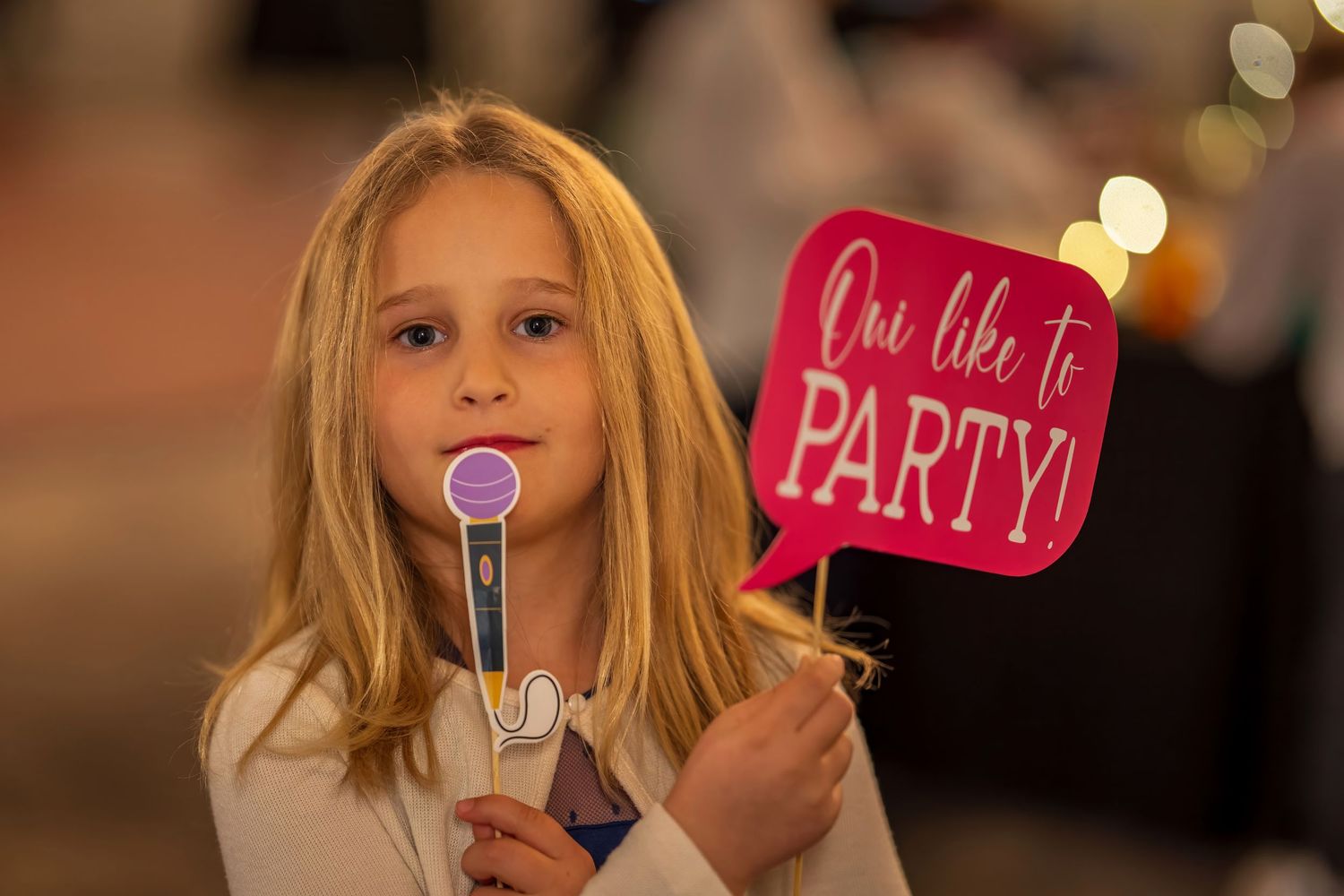 Young blonde girl holding a microphone prop and a pink party speech bubble sign at a celebration event.