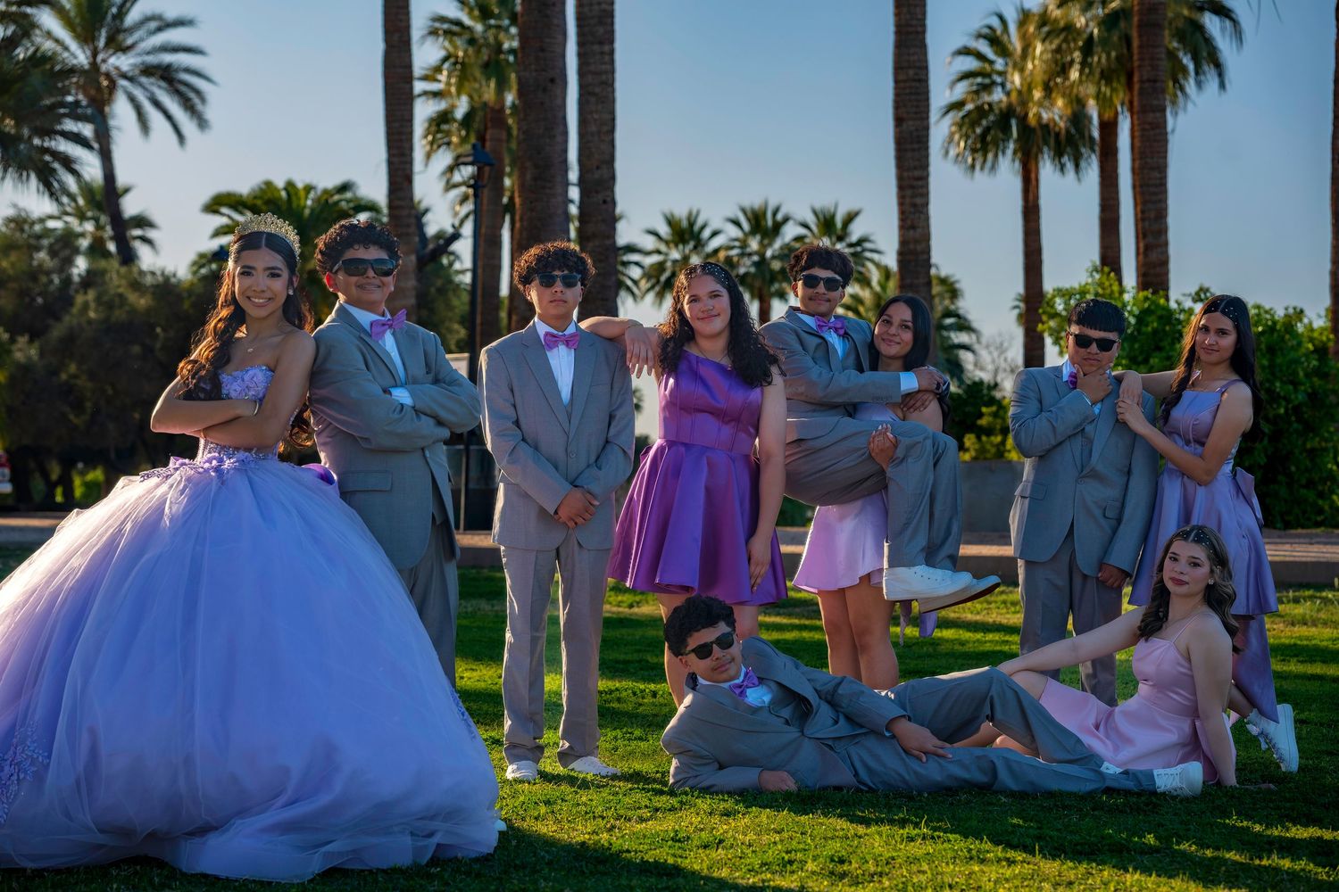 Group of teens in formal prom attire posing on green lawn with tall palm trees in background.