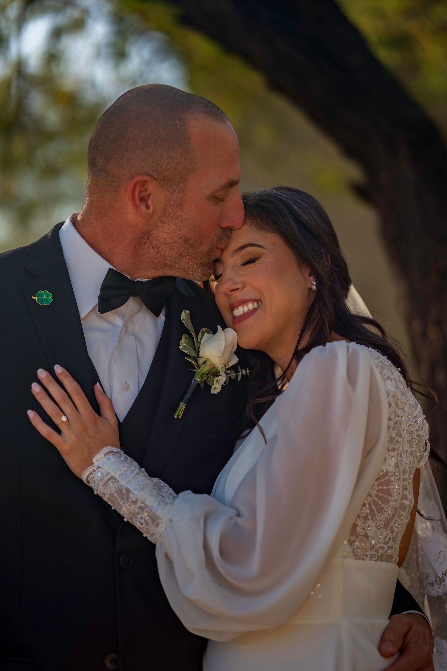 A joyful bride in a lace gown smiles as her tuxedo-clad groom tenderly kisses her forehead outdoors.