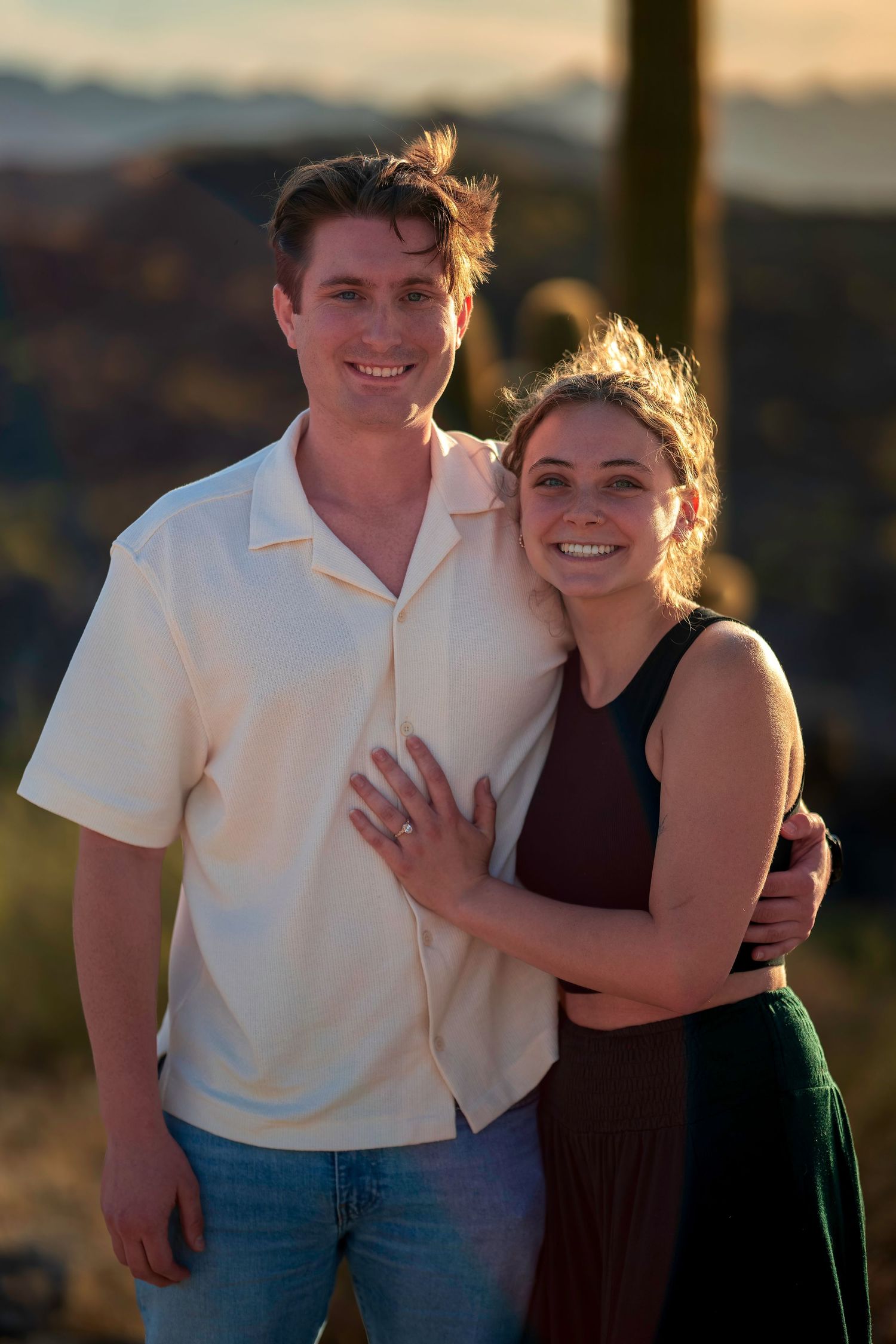 Smiling couple embracing outdoors at golden hour with saguaro cacti and desert landscape in background.