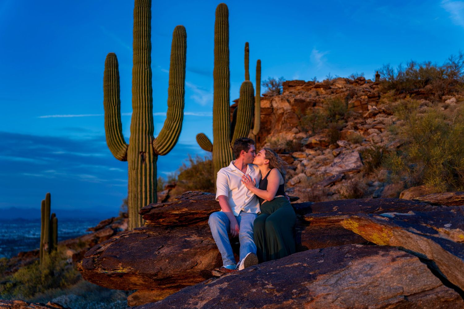 Couple sharing a romantic moment on rocks surrounded by tall saguaro cacti at dusk.