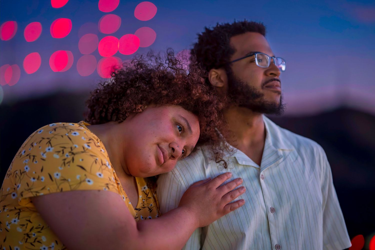 Couple leaning close together with colorful bokeh lights in background at night.