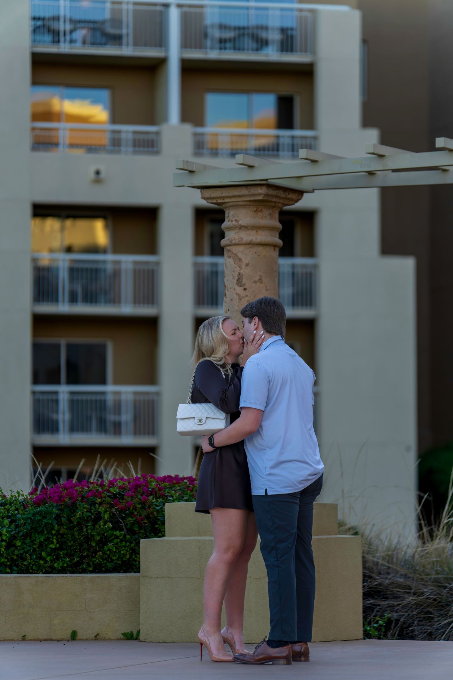 Couple sharing a romantic kiss outdoors near a resort building with balconies and colorful flowers.