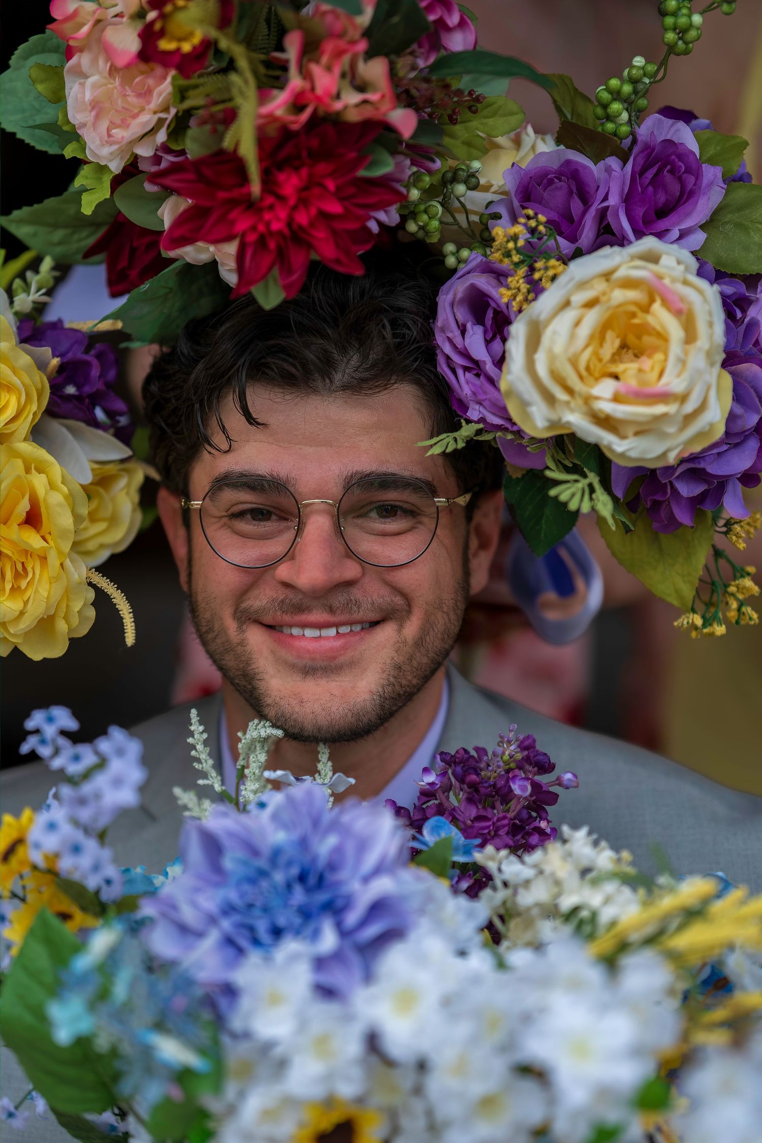 Smiling young man with glasses surrounded by colorful artificial flowers including purple, yellow, and red blooms.