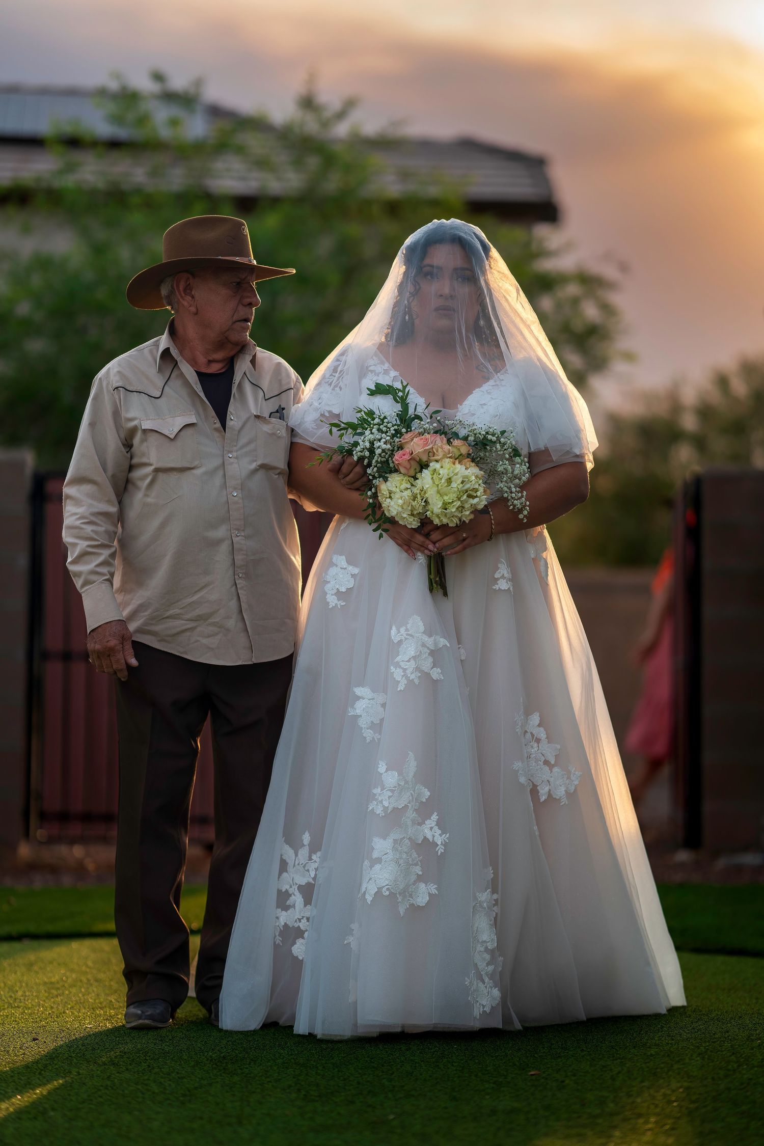 Bride in white lace gown with veil holds bouquet beside older man in cowboy hat at sunset outdoor wedding.