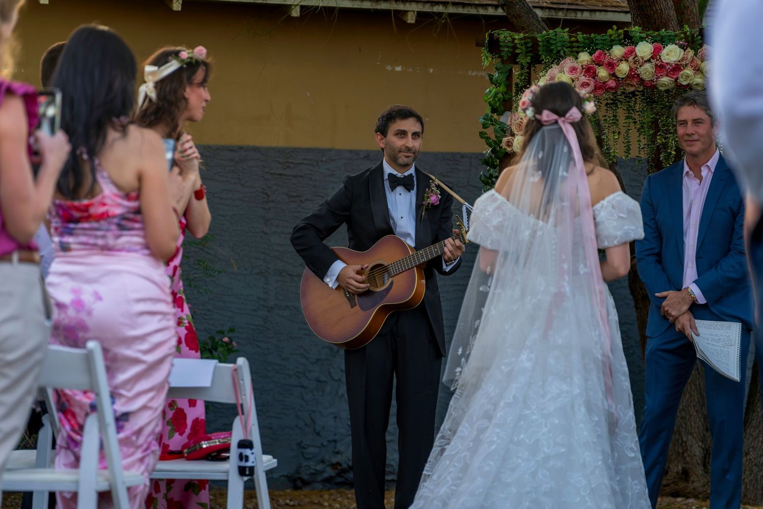 Groom plays acoustic guitar for bride at outdoor wedding ceremony with floral arch backdrop.