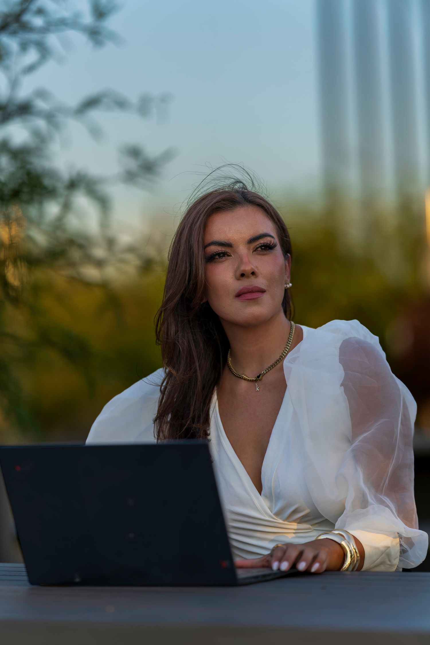 Elegant woman in white sheer blouse working on laptop outdoors at sunset.