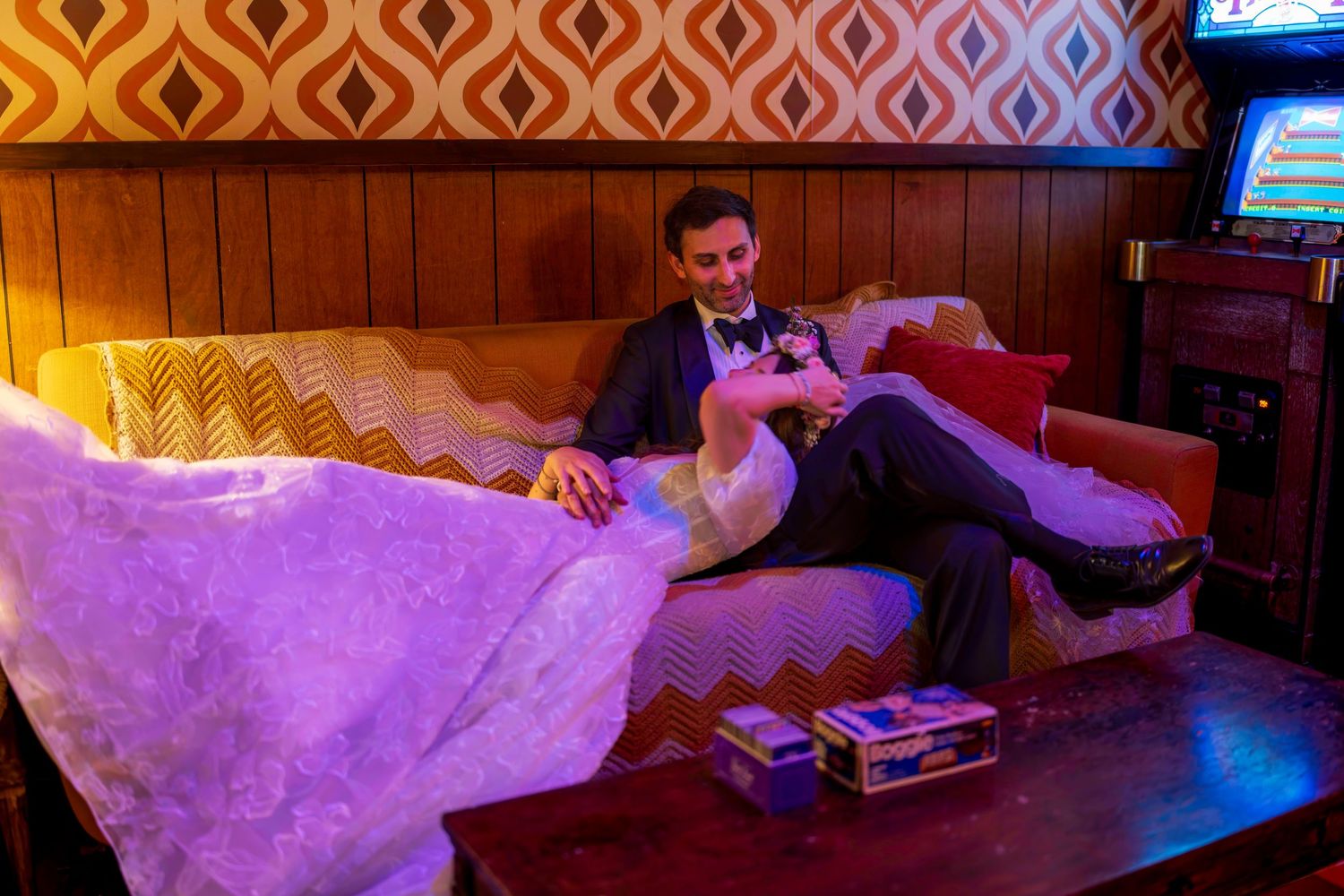 Bride and groom relaxing on a vintage couch in a retro-styled venue with warm purple lighting.