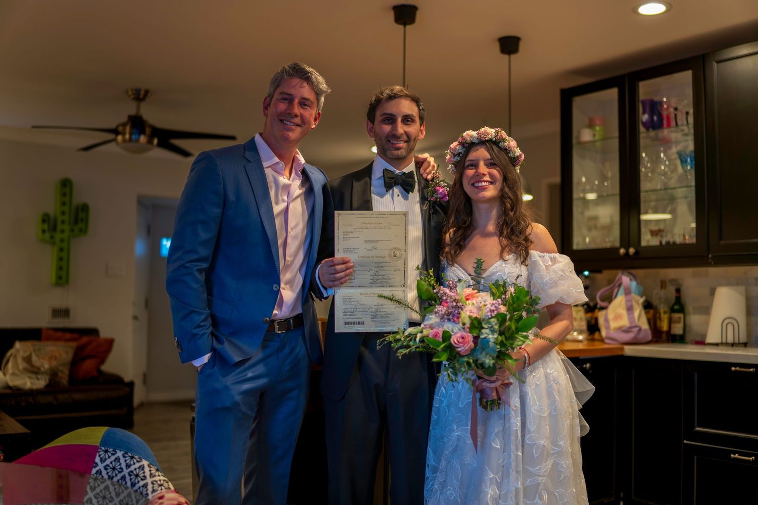 Newlywed couple holding marriage certificate with officiant in a decorated indoor wedding ceremony.