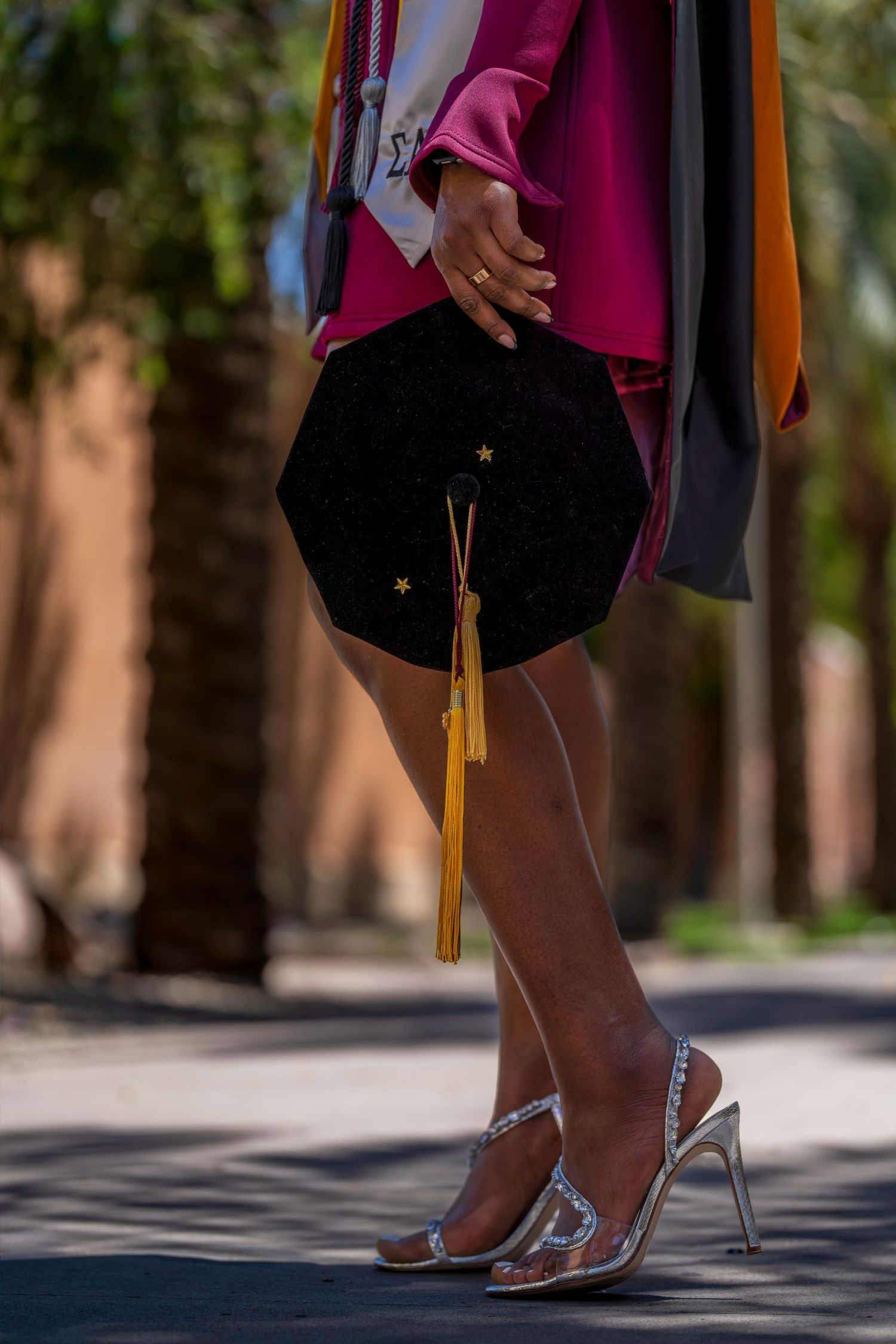 Graduate holding black cap with gold tassel, wearing silver heels and colorful academic regalia outdoors.