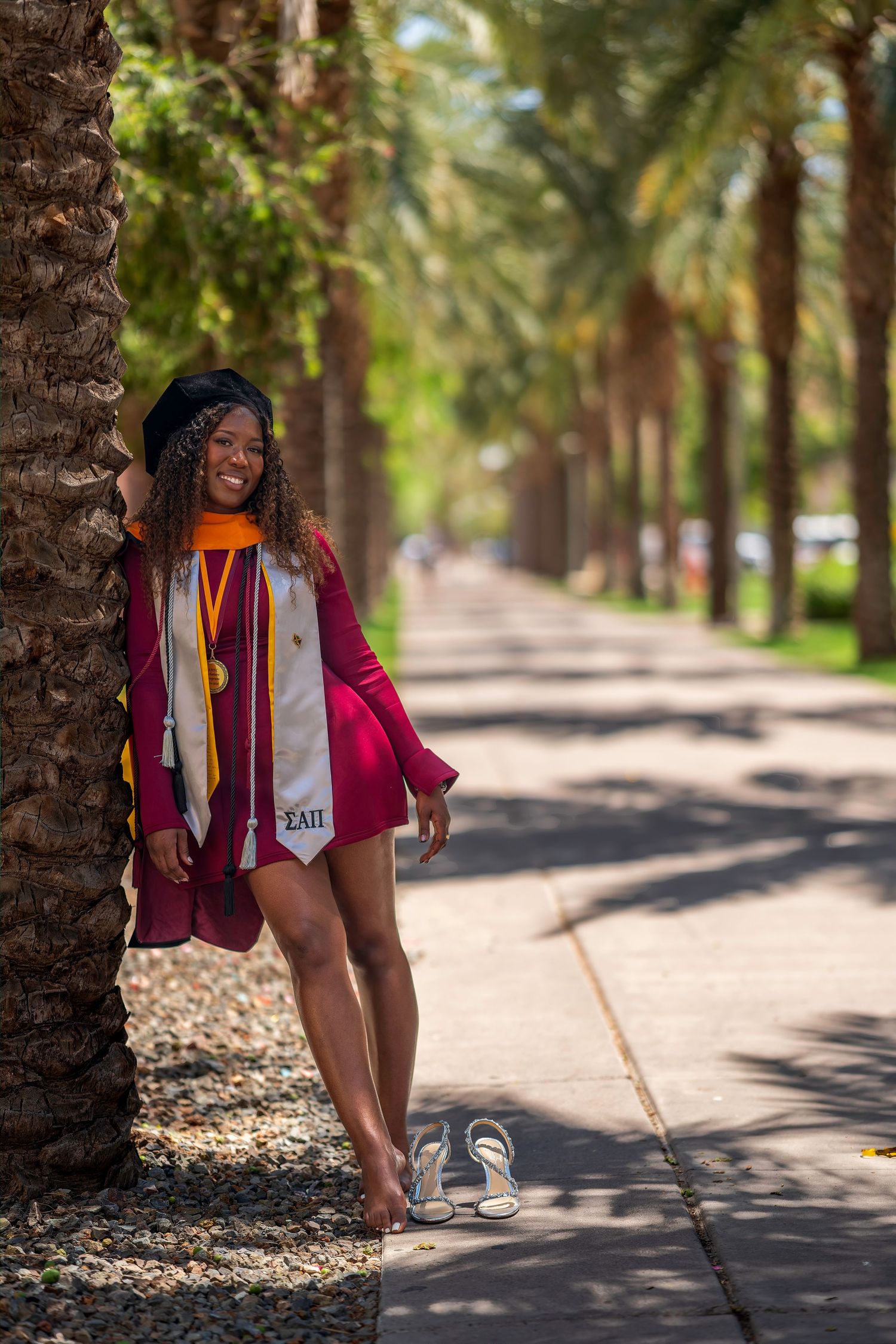 Young woman in maroon graduation gown and cap leans against palm tree on sunny campus walkway.
