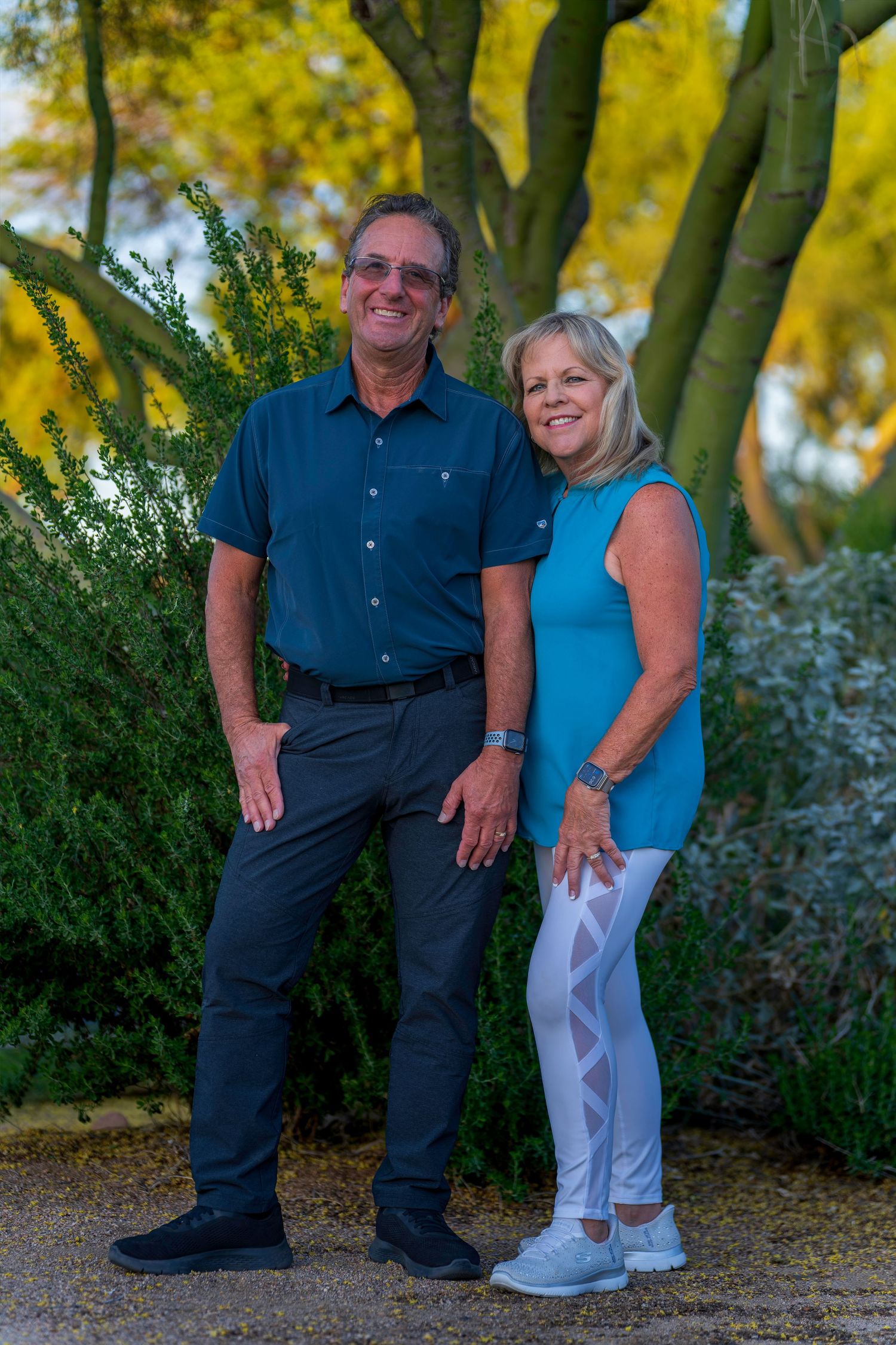 A smiling older couple poses together outdoors near green shrubs and a yellow tree in warm natural lighting.