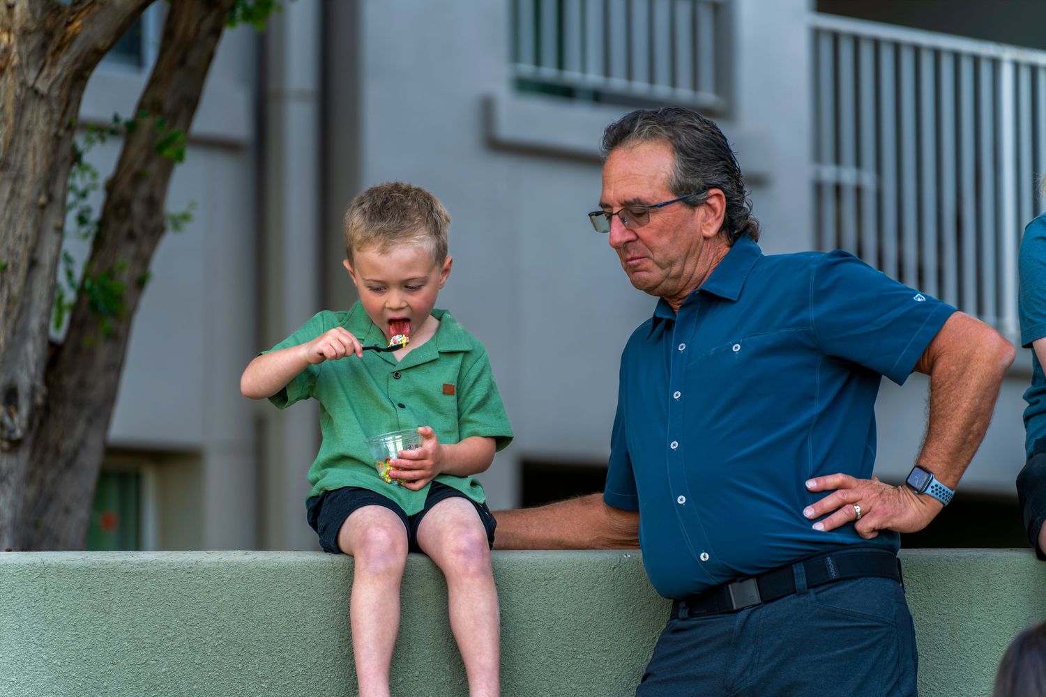 A young boy eats a snack while sitting on a wall next to an older man at a resort.