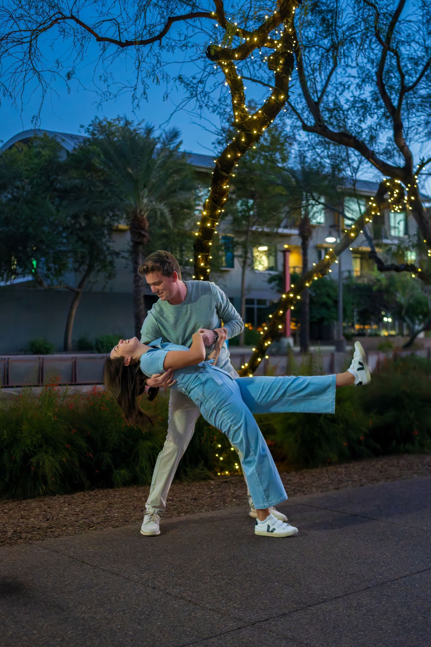 Couple dancing outdoors at night, man dipping woman under trees decorated with string lights.