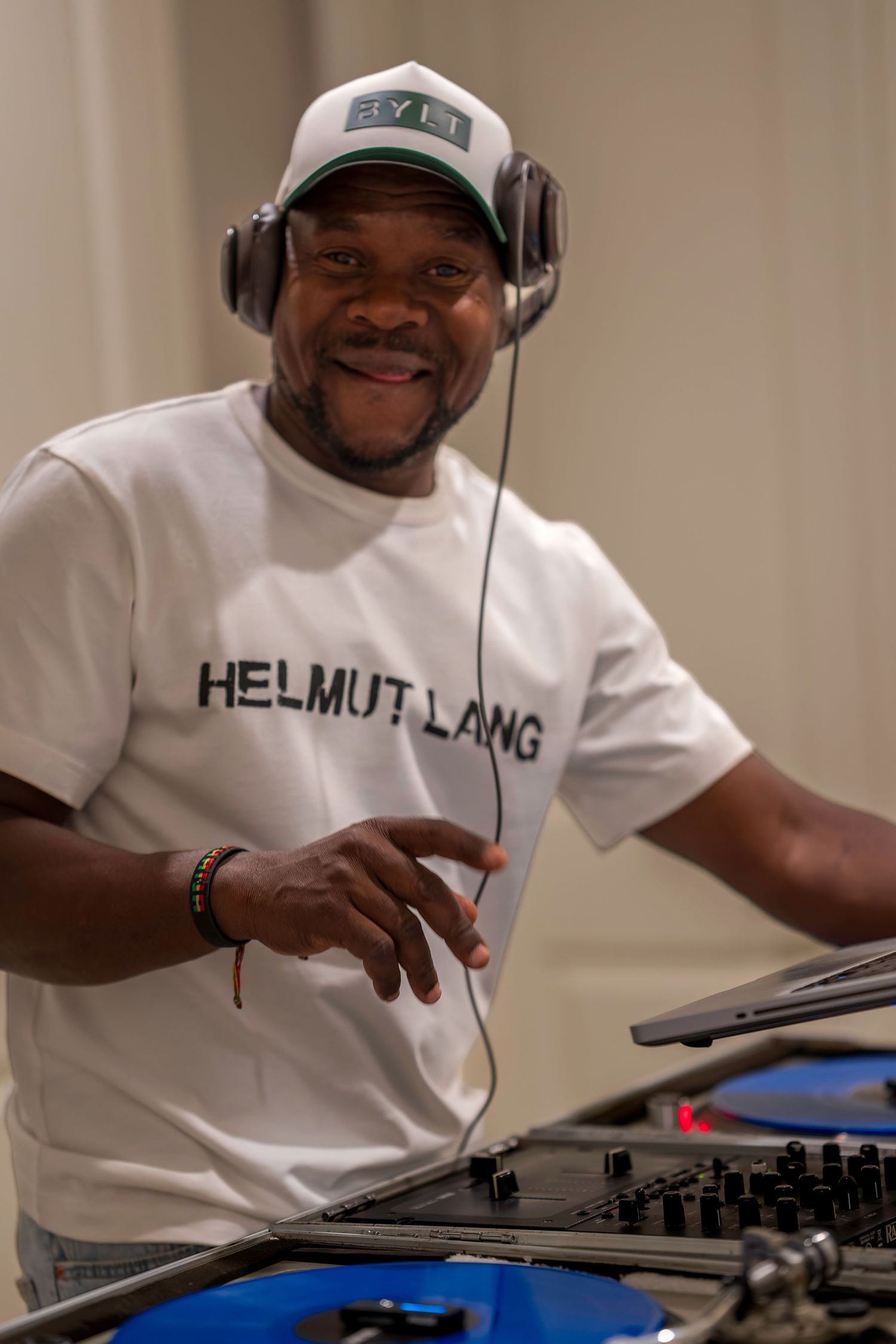 Smiling DJ wearing Helmut Lang shirt and headphones operating a mixing console at an event.