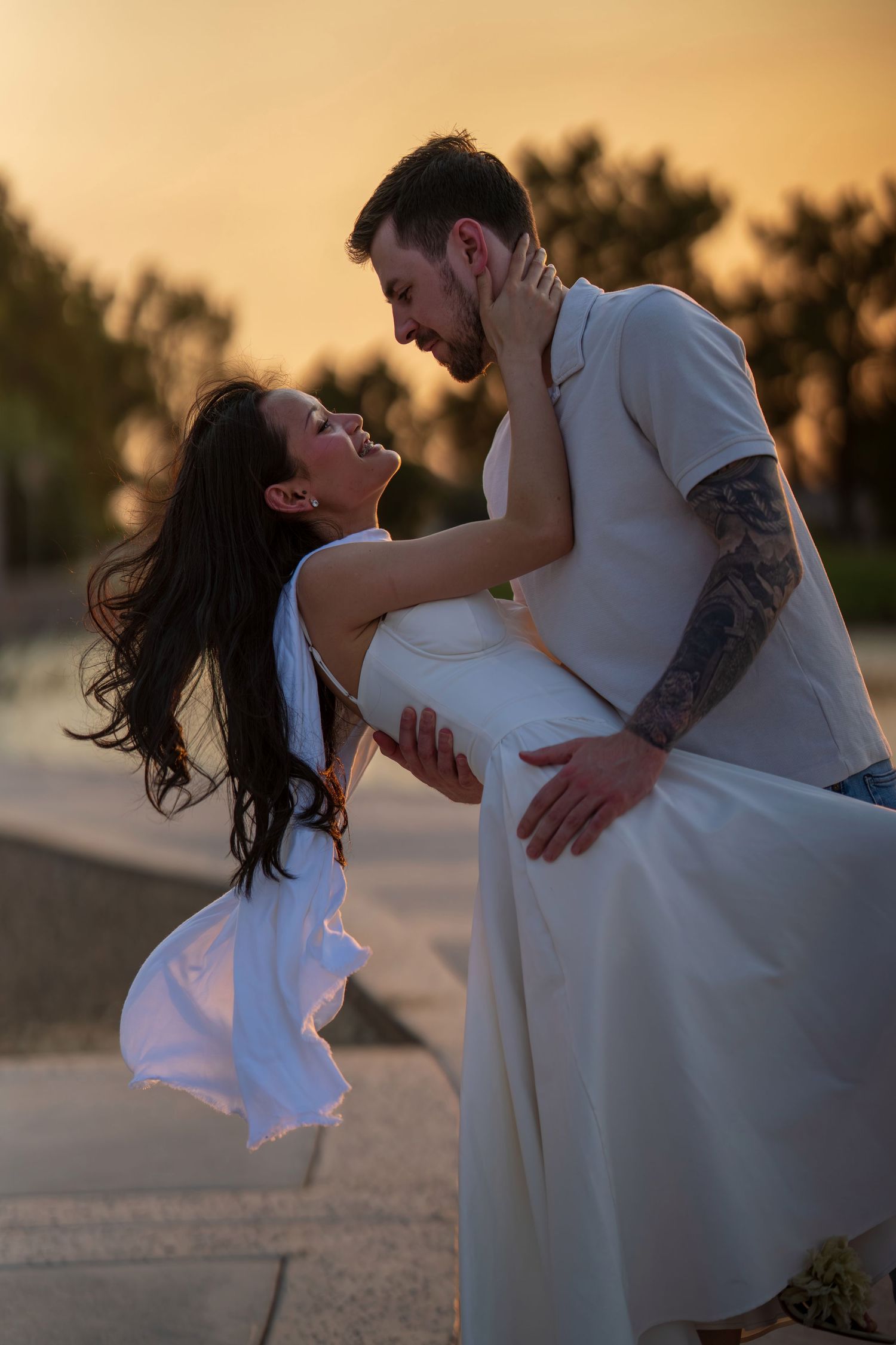 Couple sharing romantic moment at sunset, woman in white dress being dipped by tattooed man outdoors.
