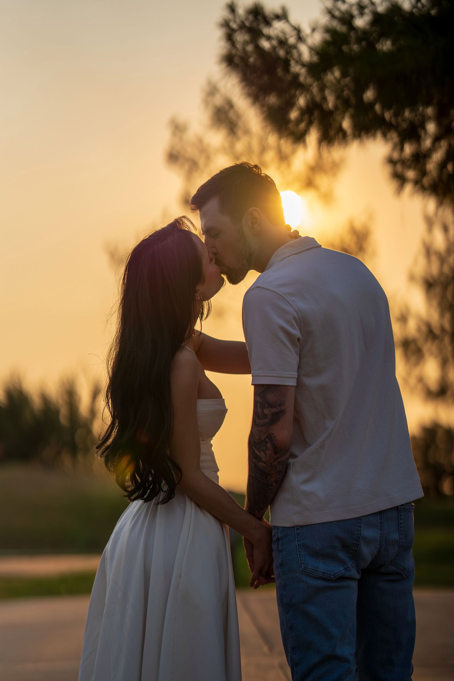Couple sharing a romantic kiss at golden sunset, woman in white dress and tattooed man in white tee holding hands outdoors.