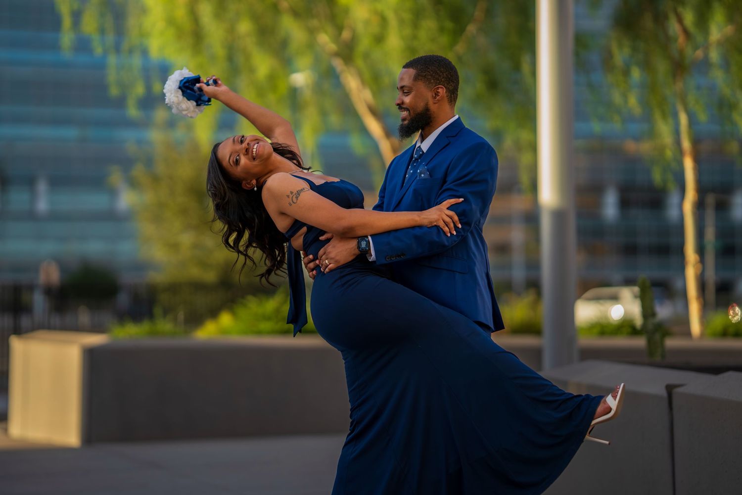 Couple in matching navy blue outfits dancing outdoors, woman in flowing gown leaning back holding flowers.