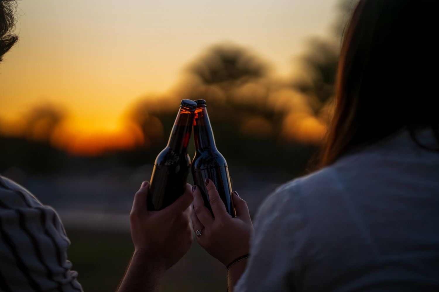 Two people clinking beer bottles together against a vibrant orange sunset sky outdoors.