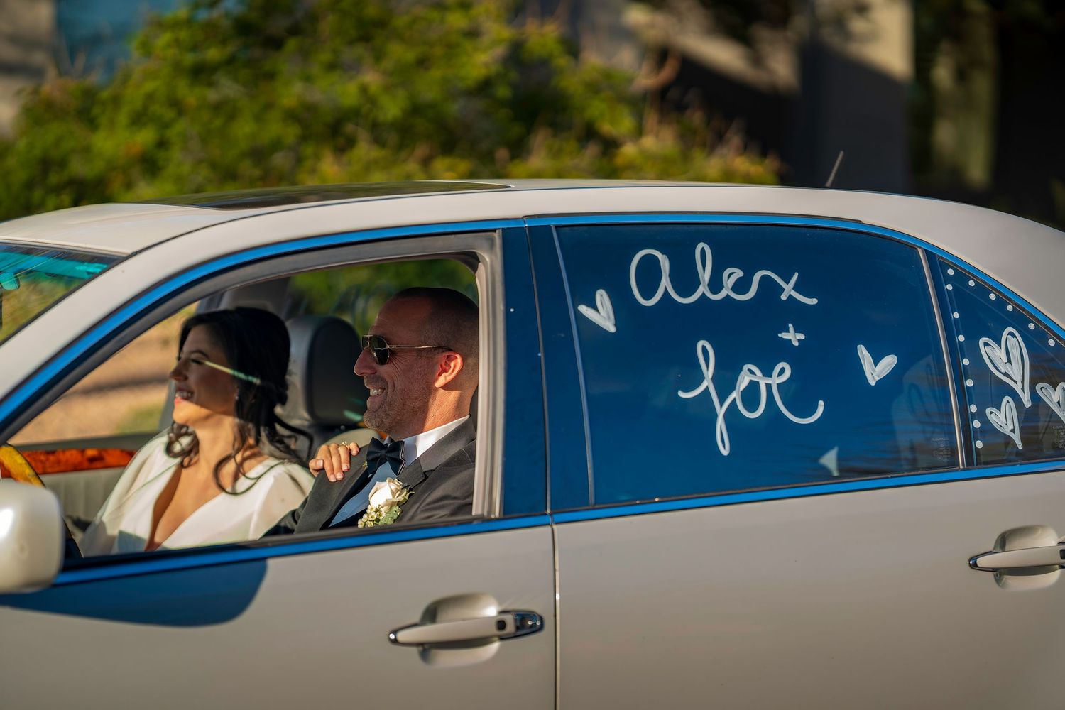 Newlyweds riding in decorated wedding car with names Alex and Joe written on window.