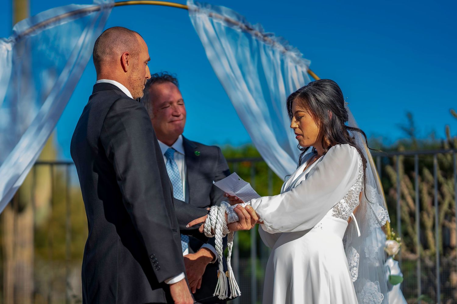 Bride and groom exchange vows under blue draped arch during outdoor wedding ceremony.