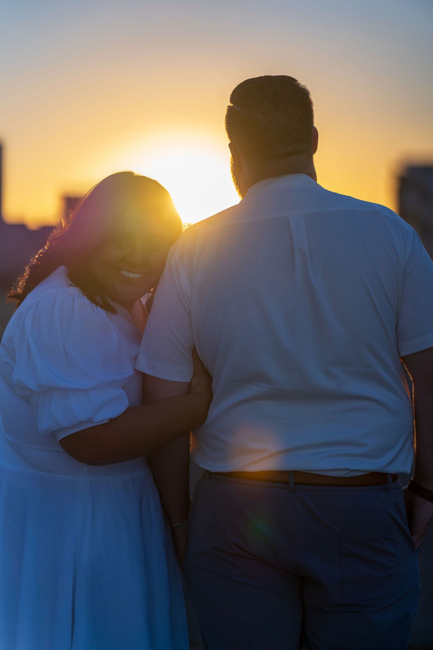 Couple embracing at sunset with golden sky, woman in white dress smiling, romantic outdoor moment backlit by warm sunlight.