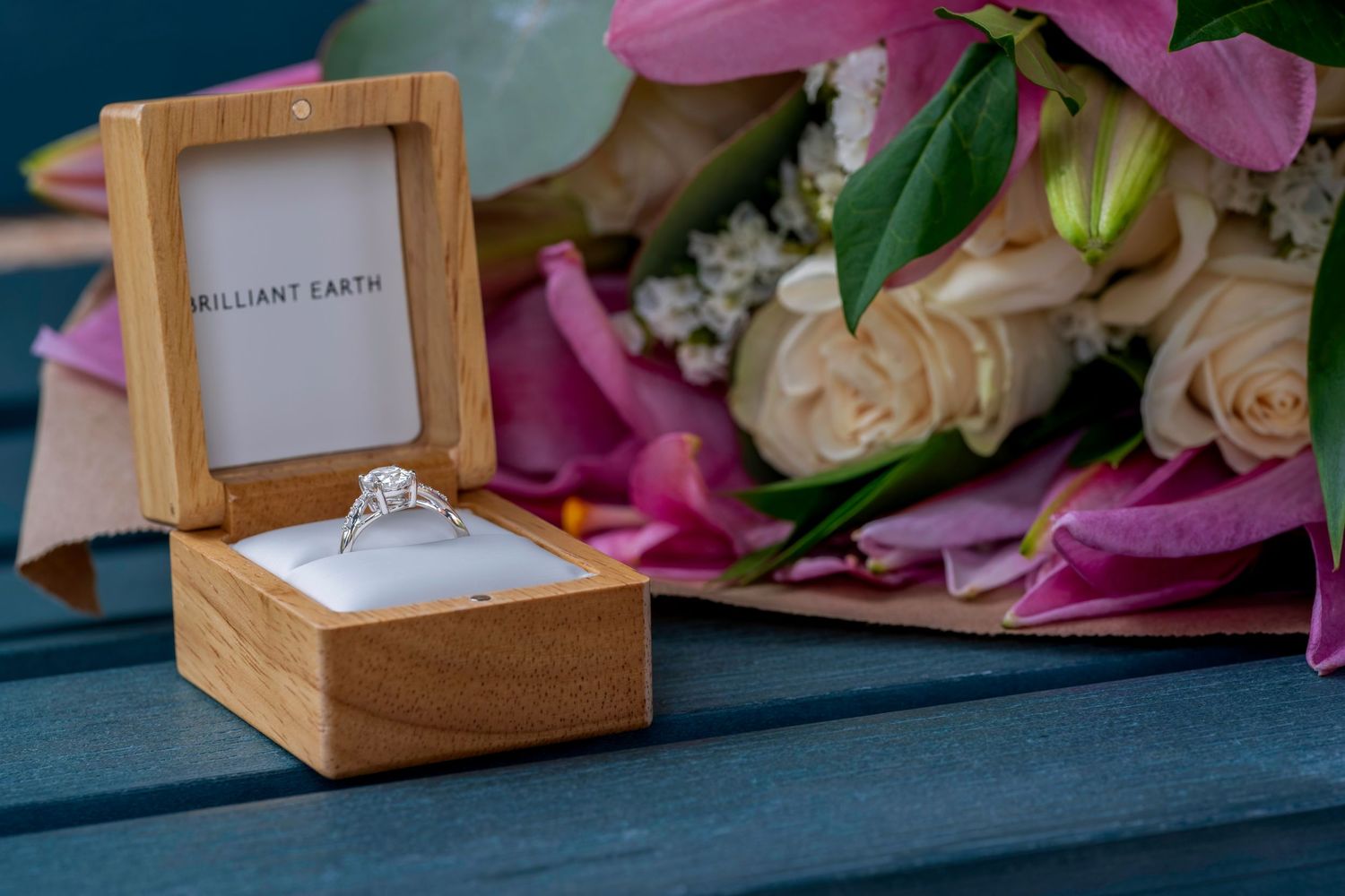 A Brilliant Earth diamond engagement ring in a wooden box beside a bouquet of pink and cream flowers.