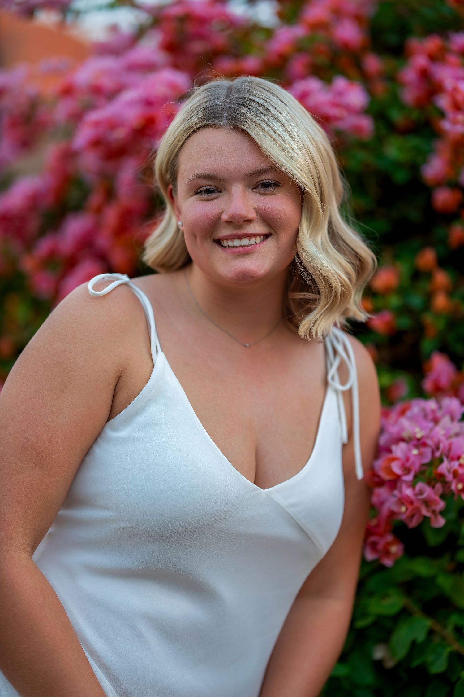 Smiling blonde woman in a white sundress posing in front of vibrant pink flowering bushes.