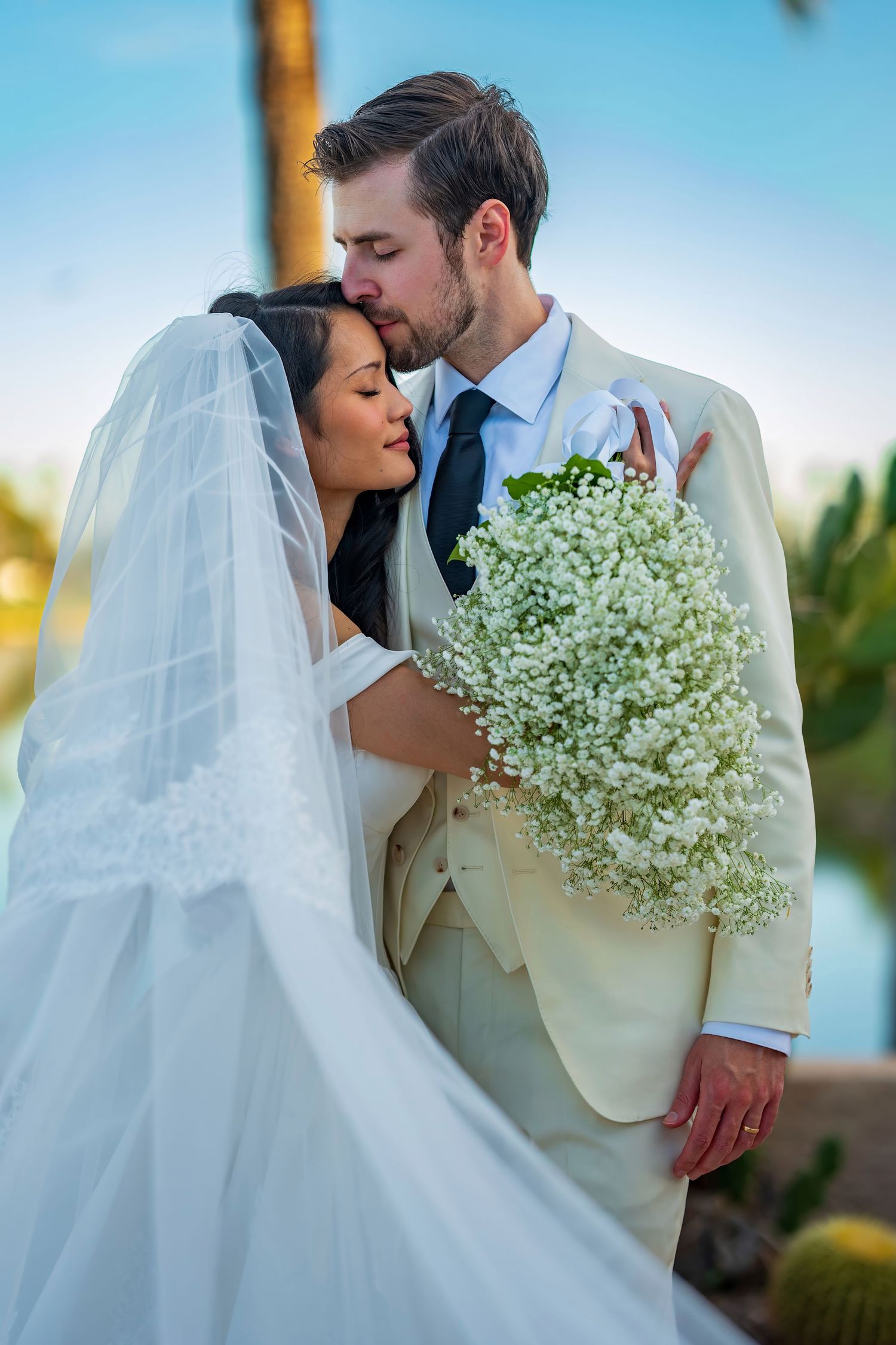 Wedding couple embracing with a bouquet of baby's breath flowers against a blue sky backdrop.