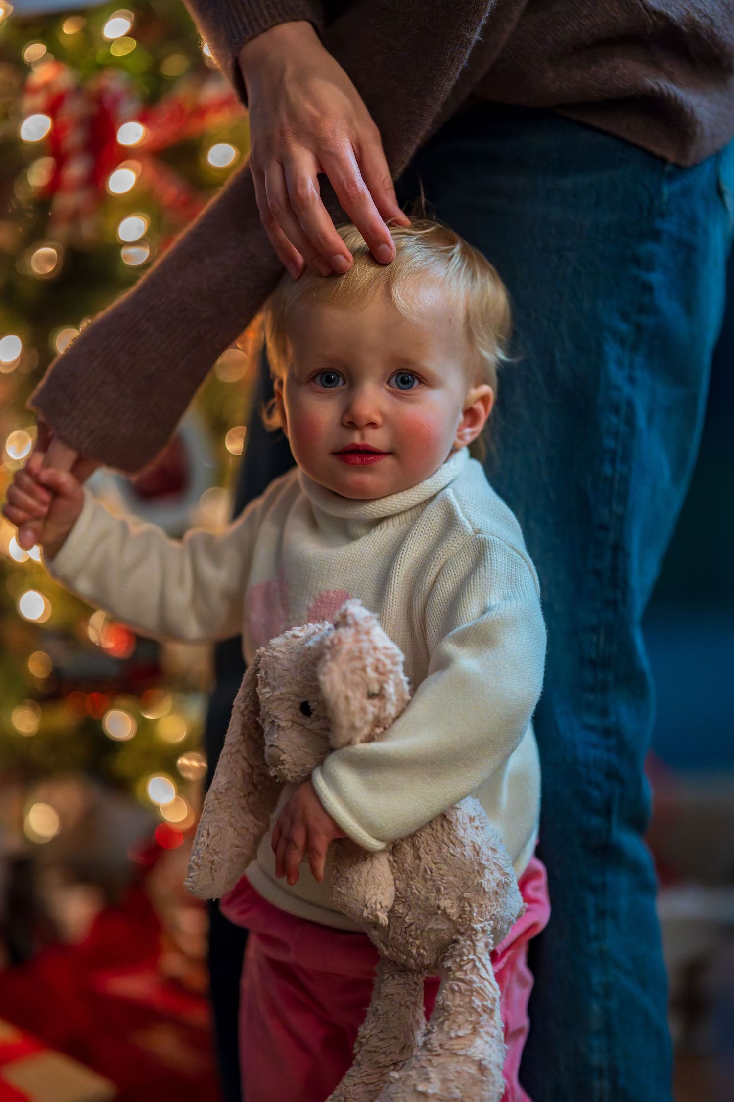 Toddler carrying stuffed animal near Christmas tree with bokeh lights.