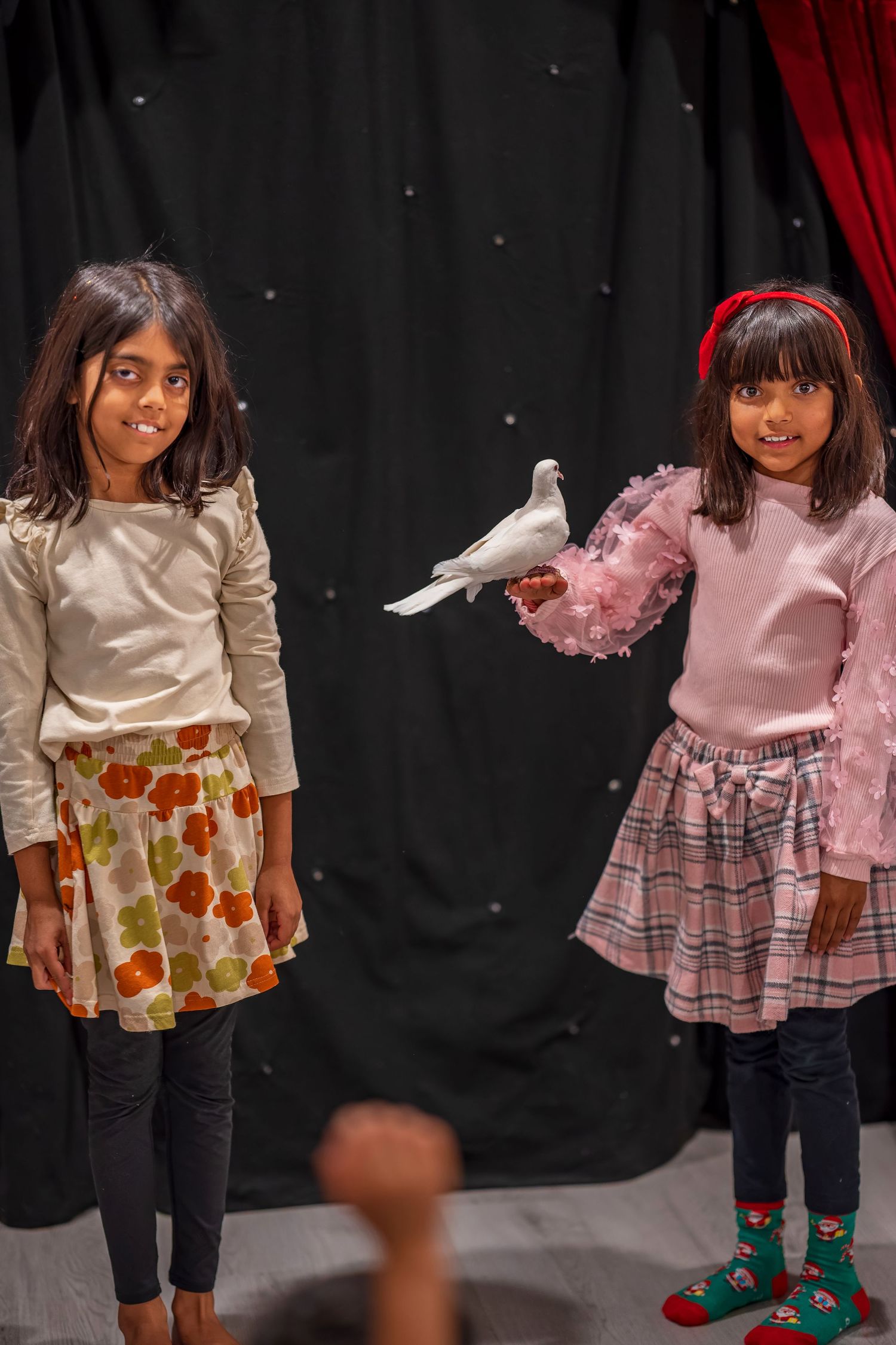 Two children in dresses interact with a white dove against a black stage backdrop.