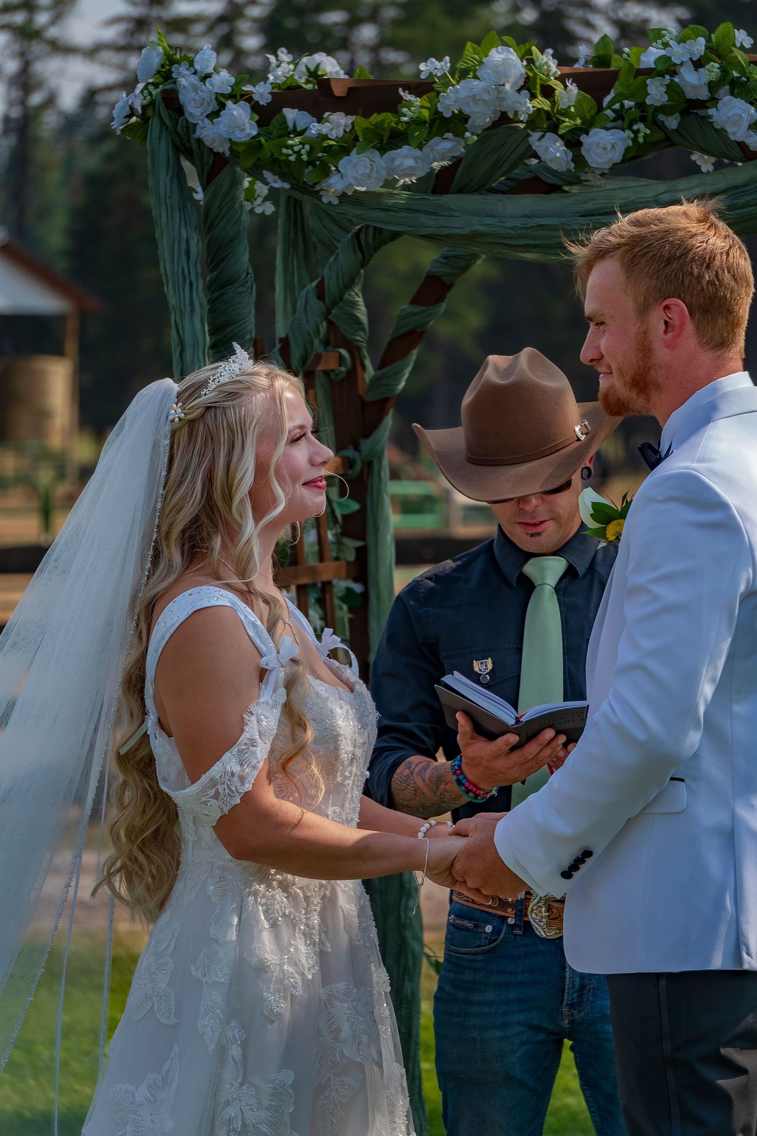 Emotional wedding ceremony moment under decorated green arbor with pine trees in background.