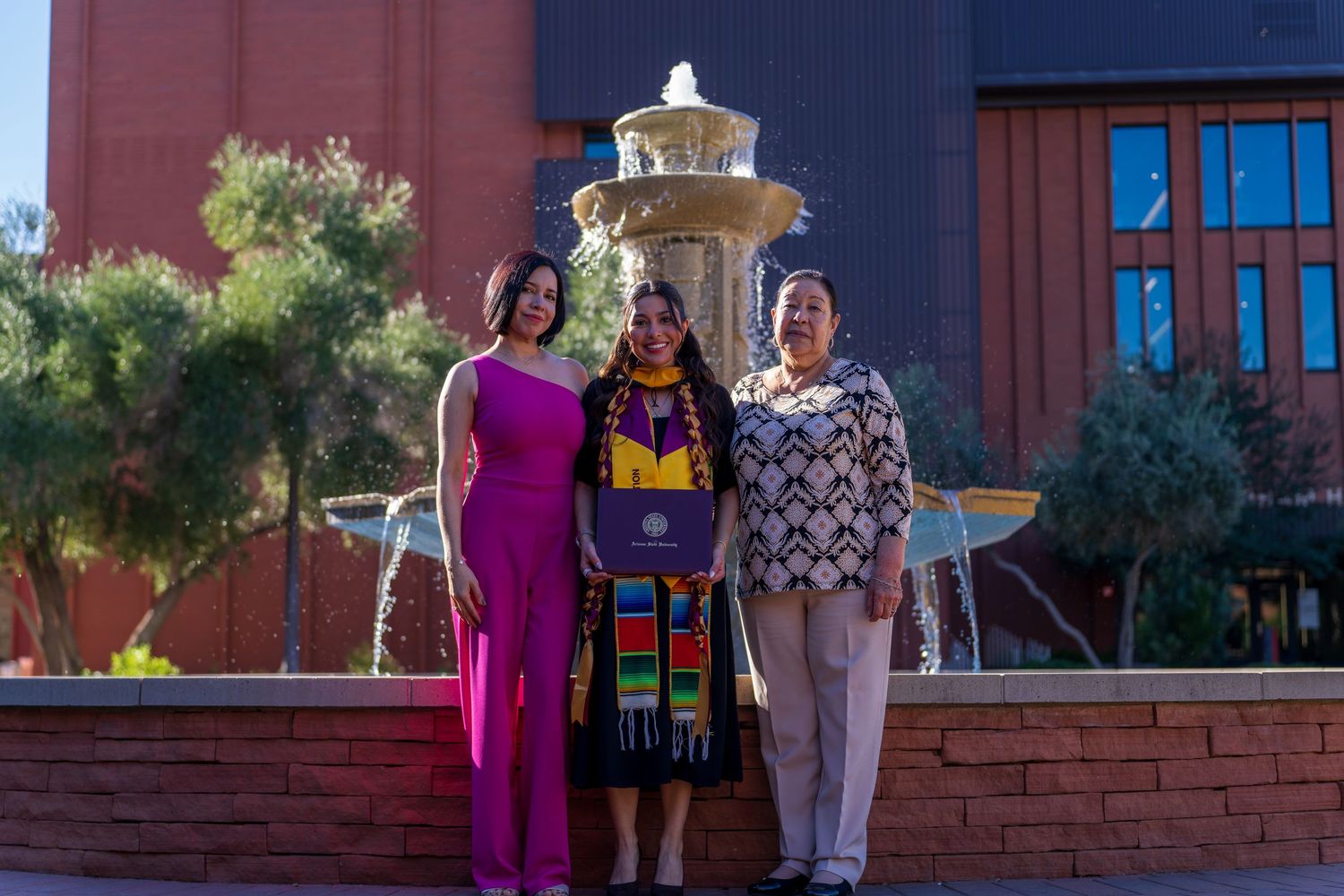Three people posing together in front of a brick building with a decorative fountain in the background.
