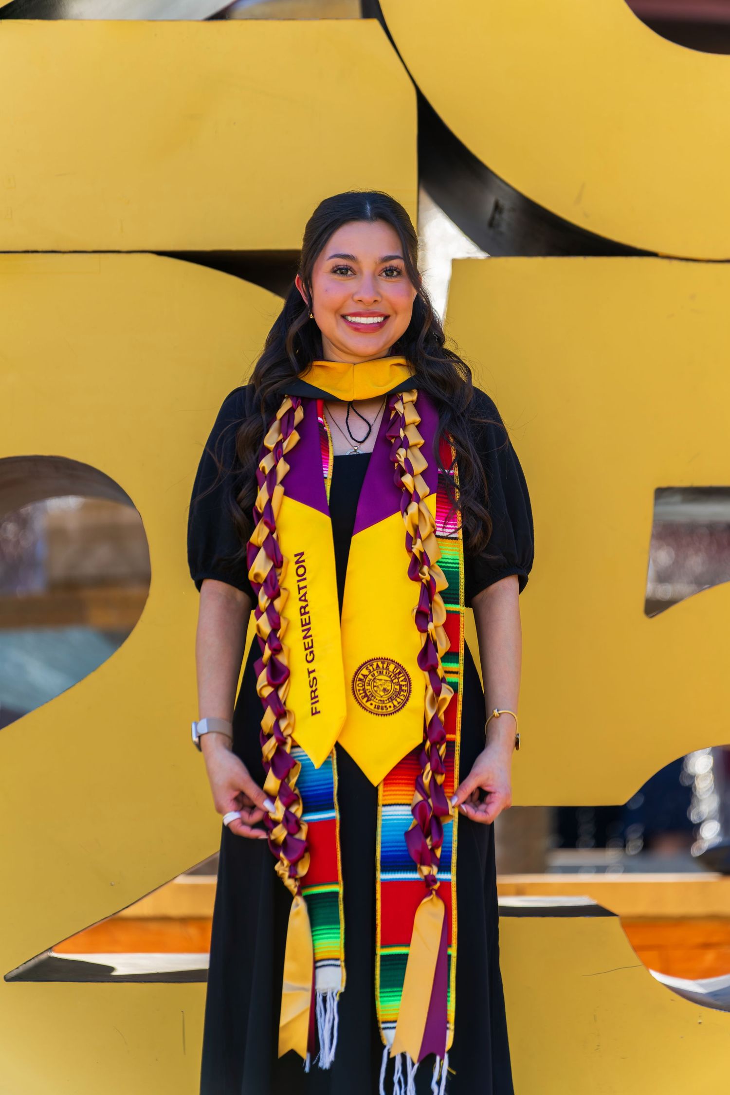Graduate in academic regalia with colorful stoles poses against a yellow geometric backdrop.