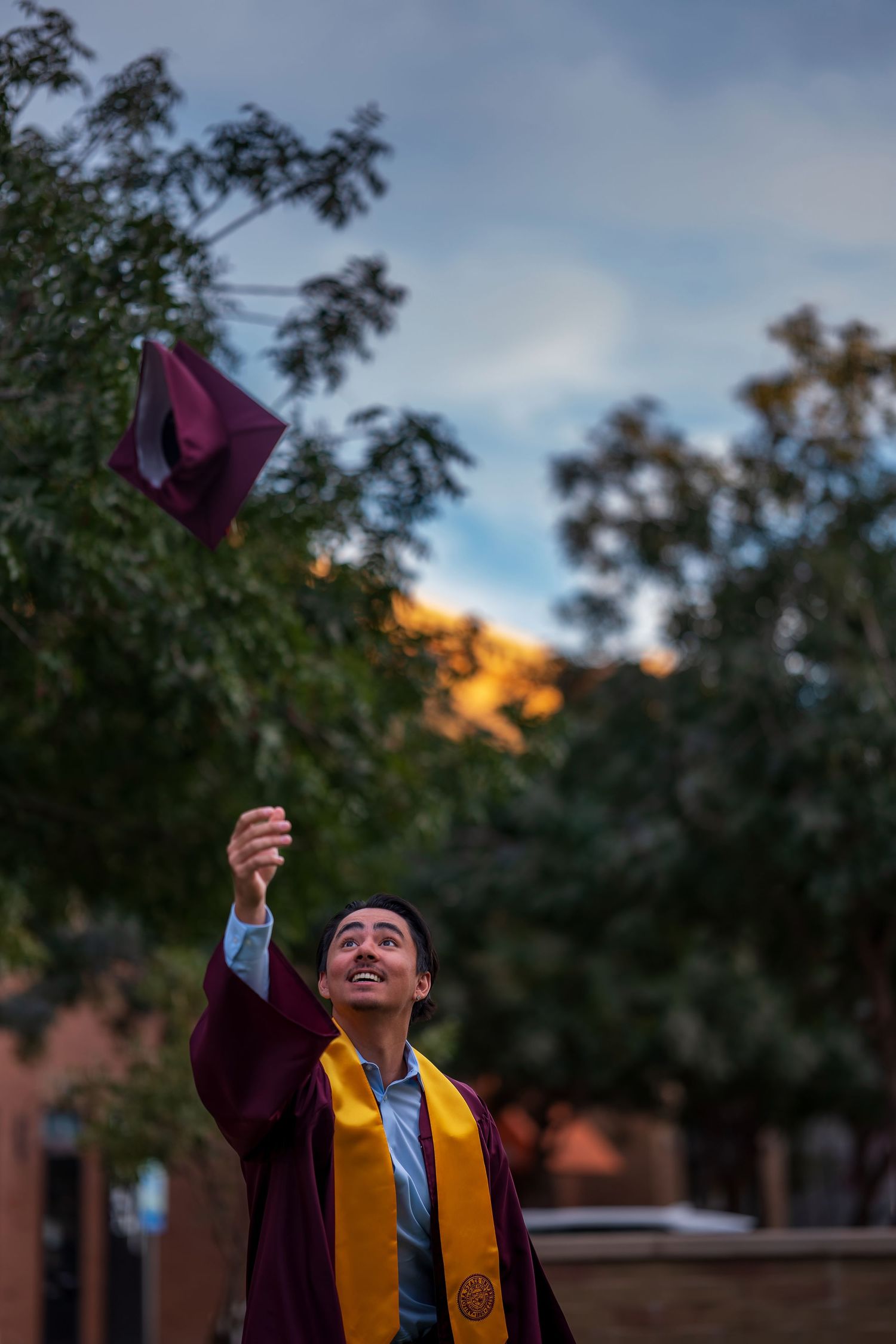 Graduate in yellow academic regalia holds up diploma against sunset sky during outdoor commencement ceremony.