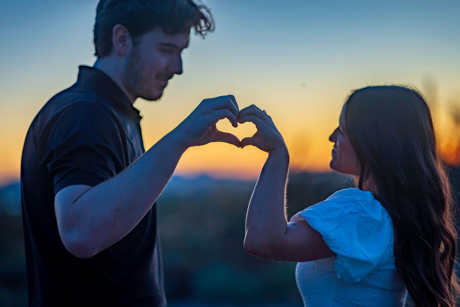 Silhouettes of a couple making heart shapes with their hands against a desert sunset.