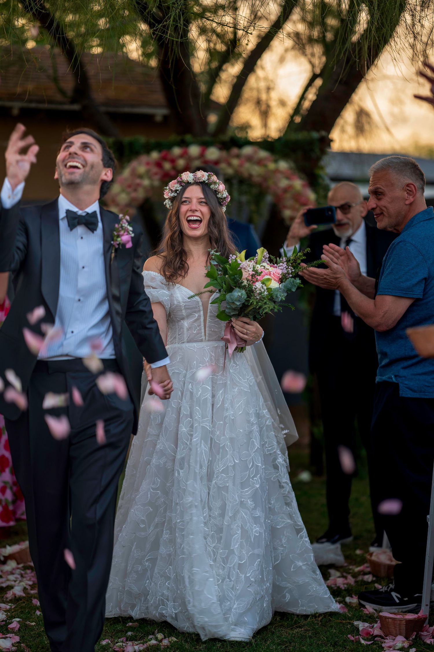 Joyful newlyweds walk down the aisle as guests toss pink flower petals at outdoor wedding ceremony.