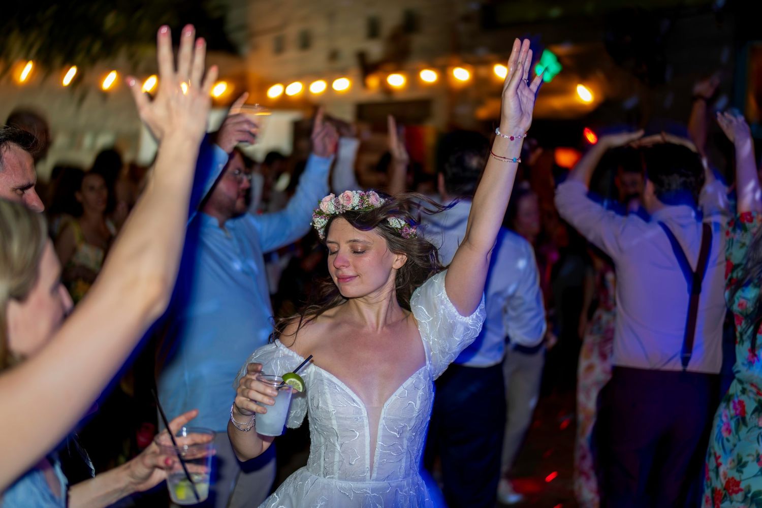 Bride in white dress and floral crown dancing joyfully with guests at an outdoor wedding reception at night.