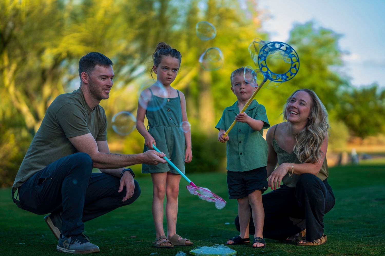 Family of four playing with giant bubbles in a lush green park at sunset.