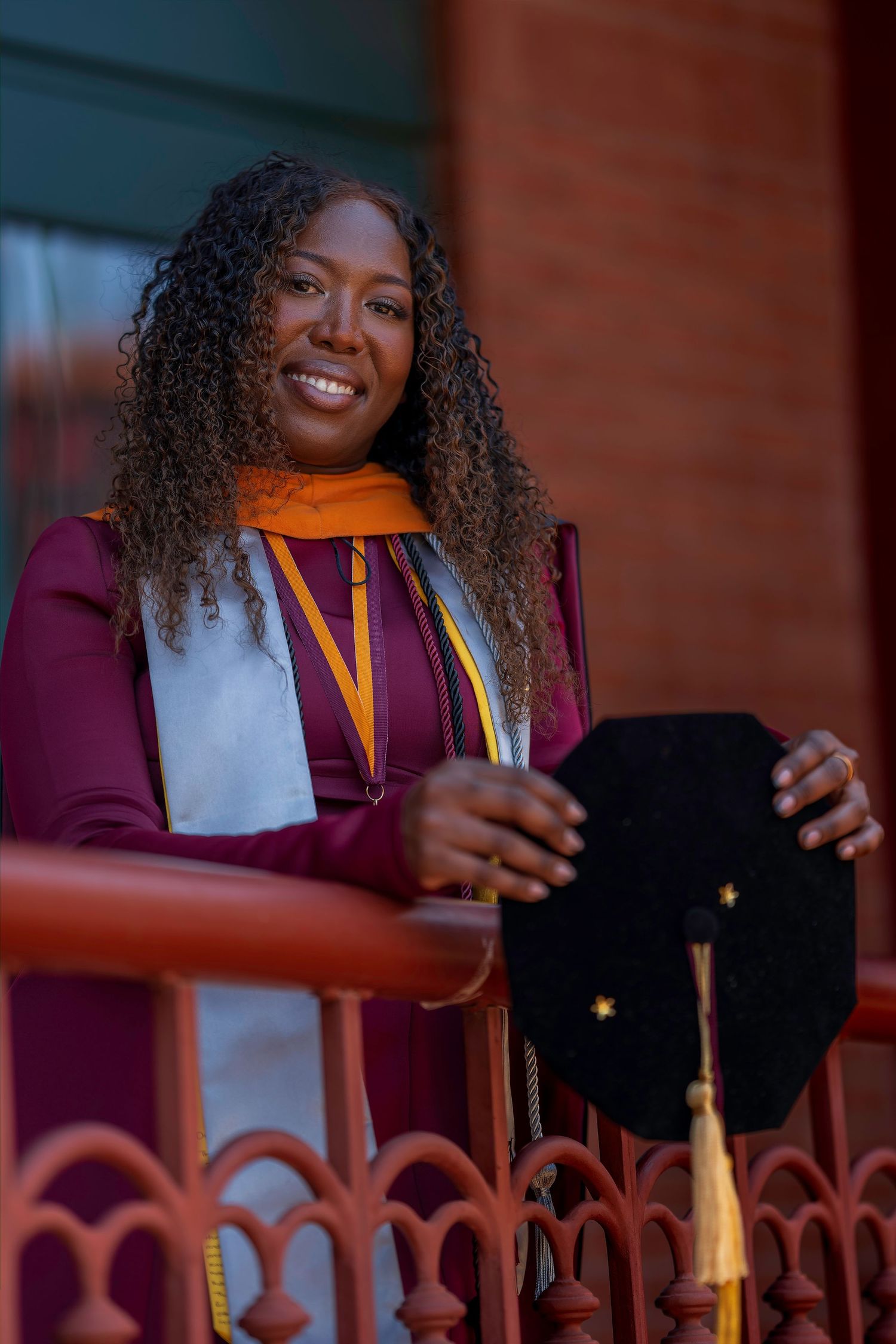 Smiling Black woman in maroon graduation gown holding cap with gold tassel, wearing honor stole on brick building balcony.
