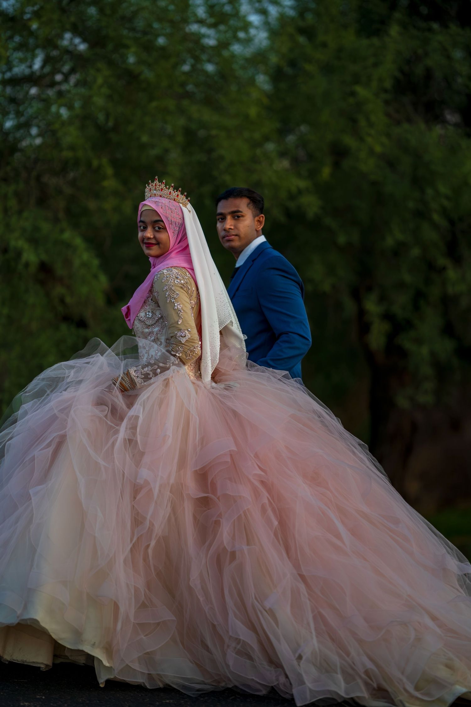 Bride in voluminous pink ball gown with hijab and crown poses back-to-back with groom in blue suit outdoors.